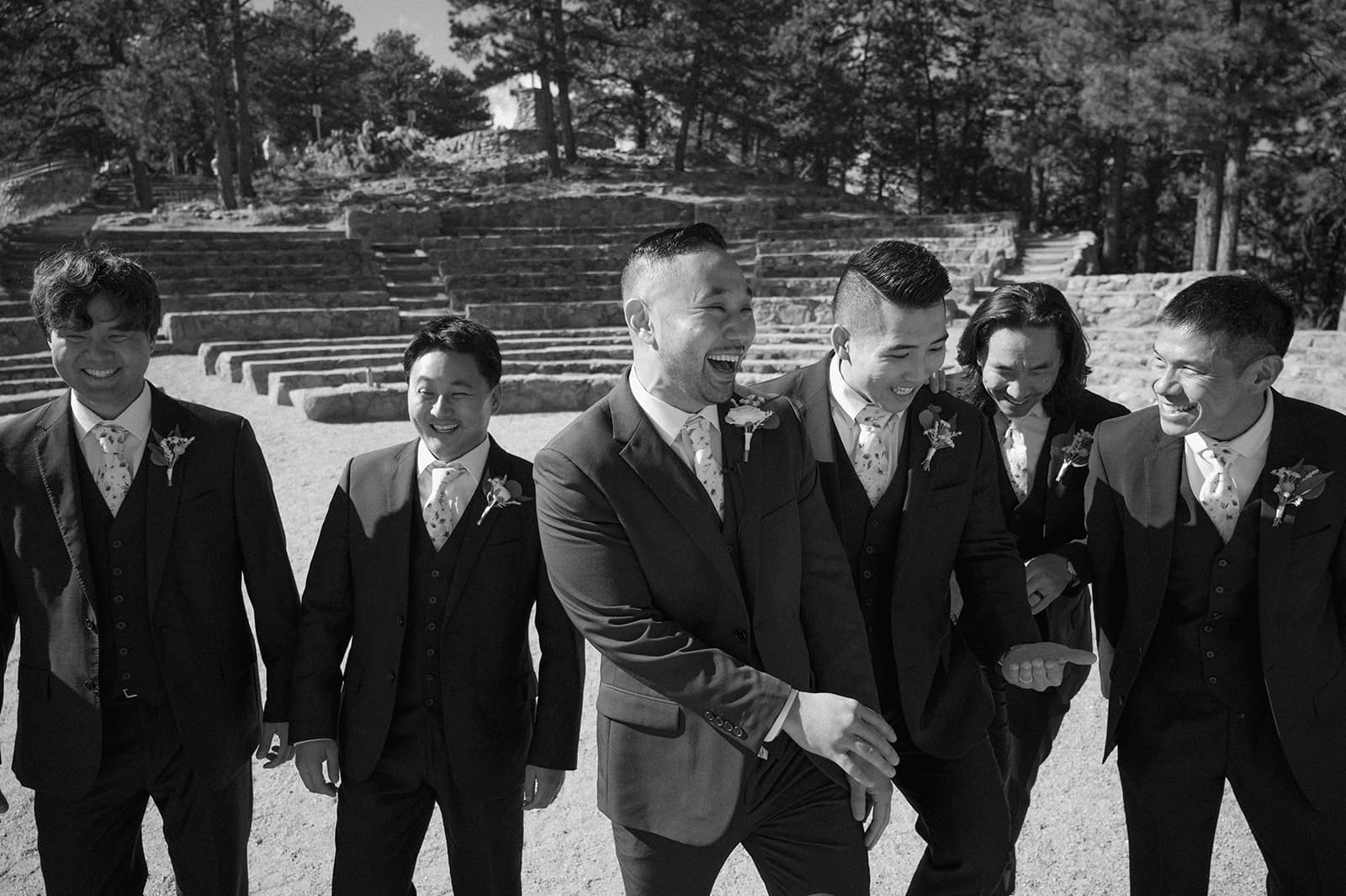 a black and white photo of a groom and his 5 groomsmen laughing as they walk towards the camera at Sunrise Ampitheater in Boulder.