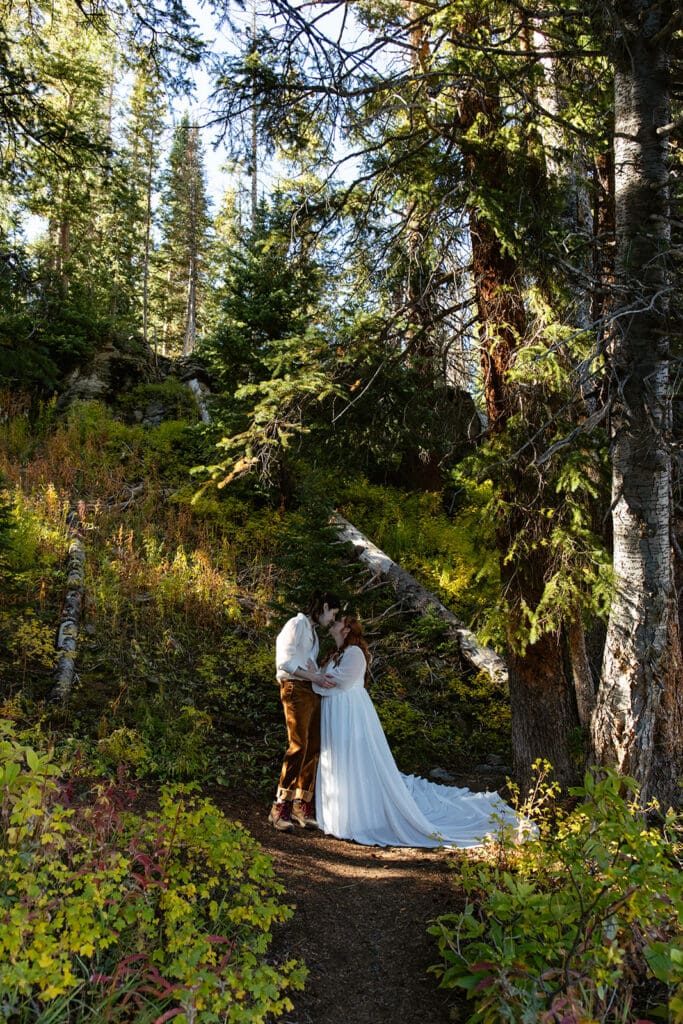 Couple stops for a kiss while hiking up a mountain trail together during their Crested Butte elopement, surrounded by tall pines and golden afternoon light.