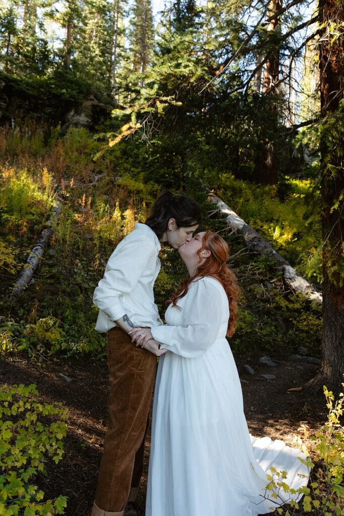 Couple sharing a kiss beneath towering evergreen trees during their Crested Butte elopement, framed by natural forest light and rich green tones.