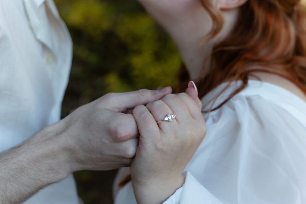Close up on a bride's ring while the groom is holding her hand. The background is blurry but you can tell they're going in for a kiss in a lush forest.