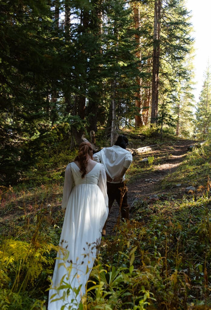 Couple hiking up a mountain trail together during their Crested Butte elopement, surrounded by tall pines and golden afternoon light.