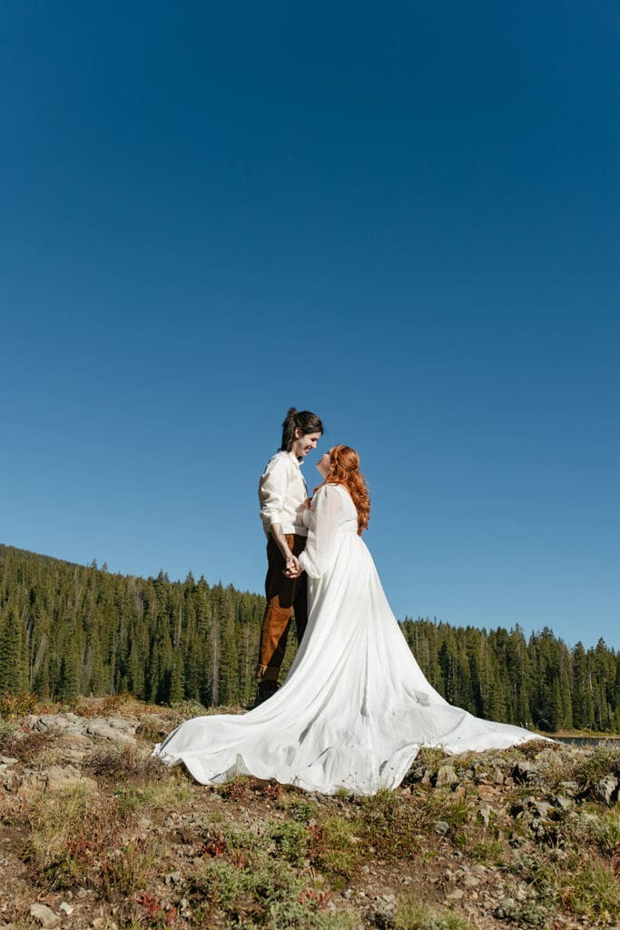 Bride and groom standing on a rocky overlook during their Crested Butte elopement, her dress flowing dramatically in the wind against a clear blue Colorado sky.
