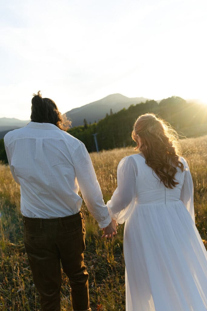 Couple walking hand in hand through a sunlit mountain meadow during their Crested Butte elopement, with rolling hills and soft golden light behind them.