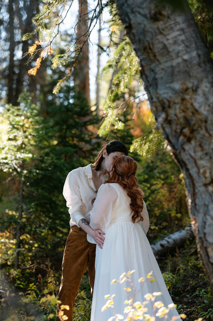 Couple sharing a kiss beneath towering evergreen trees during their Crested Butte elopement, framed by natural forest light and rich green tones.