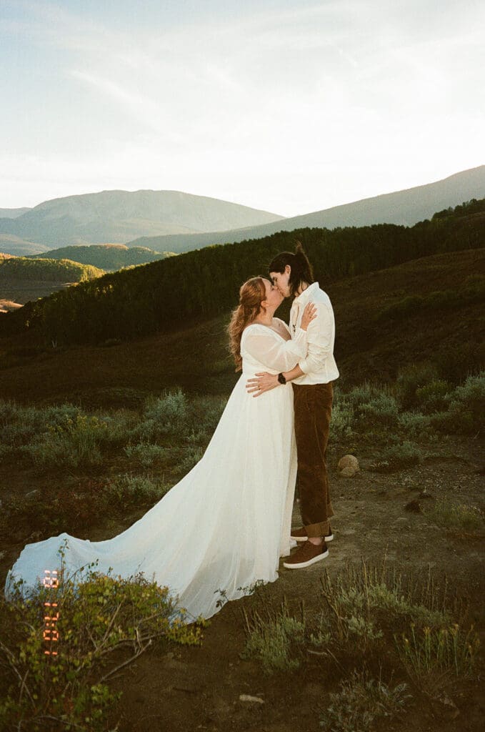 A flash film photo of a bride and groom kissing after their vows at Snodgrass Trailhead in Crested Butte with layers of lush mountains behind them.