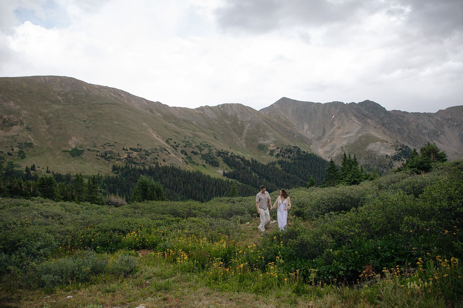 a couple in neutral, earth toned out fits walk through a field of alpine grass and wildflowers at pass lake with sweeping mountain views behind them