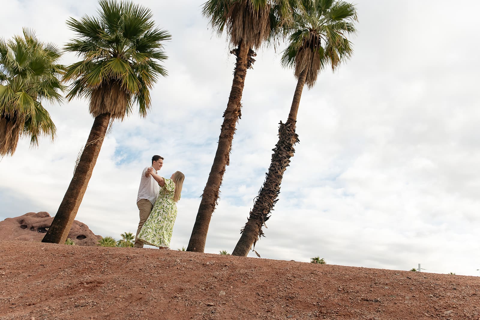a couple stands on a red dirt hill between a few palm trees dancing