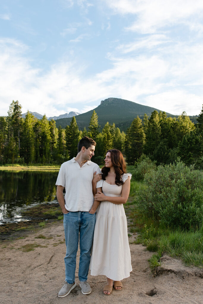 a couple stand together side by side, her arms hugging his, with the mountains and lily lake behind them.
