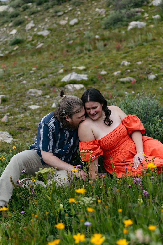 her fiance kisses her shoulder as they sit in a field of wildflowers at Pass Lake
